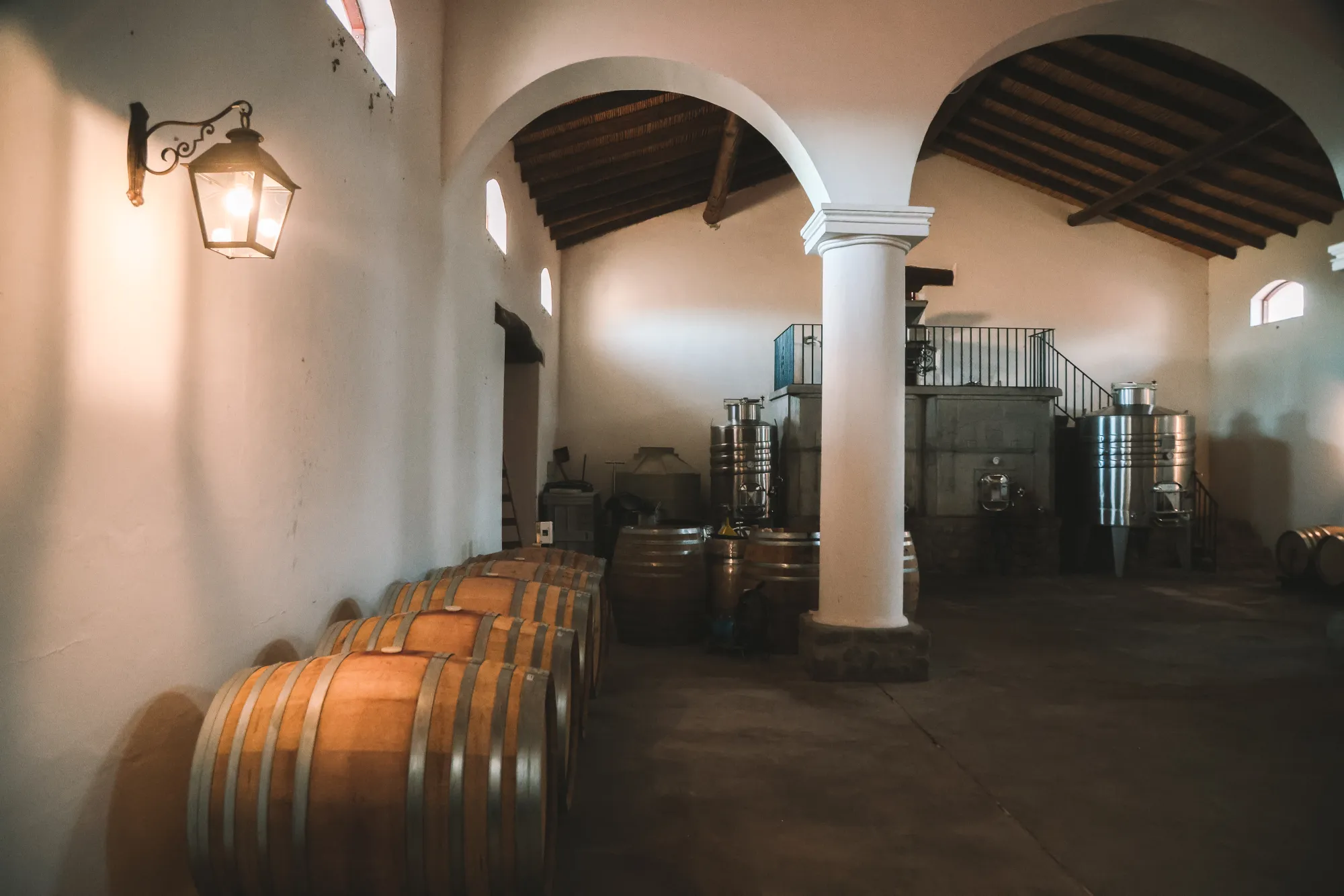 Interior cálido de la bodega con barricas, arcos blancos y farol antiguo